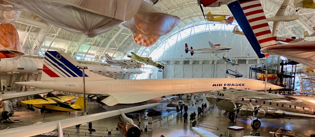 A large aviation museum exhibit featuring multiple aircraft. The main focus is a Concorde jet with Air France livery, surrounded by other historic airplanes both on the ground and suspended from the ceiling. The setting includes high ceilings with natural light streaming in through large windows.
