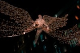 A glowing angelic figure made of intricate lights is suspended in the air, with large, luminous wings spread wide. The figure appears against a dark background, possibly in a city setting, suggested by blurred architectural elements and additional decorative lights in the background.