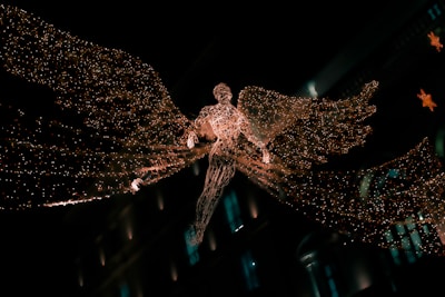 A glowing angelic figure made of intricate lights is suspended in the air, with large, luminous wings spread wide. The figure appears against a dark background, possibly in a city setting, suggested by blurred architectural elements and additional decorative lights in the background.