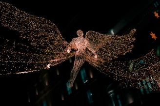 A glowing angelic figure made of intricate lights is suspended in the air, with large, luminous wings spread wide. The figure appears against a dark background, possibly in a city setting, suggested by blurred architectural elements and additional decorative lights in the background.