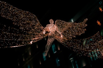 A glowing angelic figure made of intricate lights is suspended in the air, with large, luminous wings spread wide. The figure appears against a dark background, possibly in a city setting, suggested by blurred architectural elements and additional decorative lights in the background.