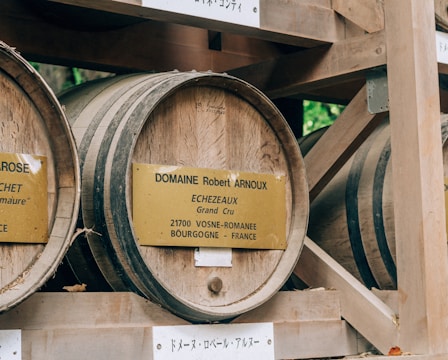 Several large wooden barrels are stored in a wooden rack structure. Each barrel has a golden plaque with French text, indicating details about the contents or provenance of the barrels. The environment appears rustic and outdoors, suggested by the hint of greenery in the background.