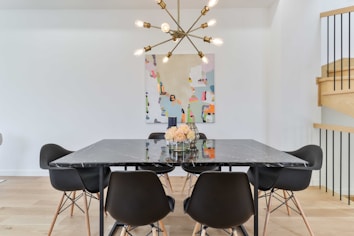 A modern dining room setting featuring a black marble table with six black chairs around it. A vase with flowers sits on the table, and above it hangs a stylish, geometric chandelier with exposed bulbs. A colorful abstract painting decorates the white wall behind the table, and wooden stairs with black railings are visible to the right.