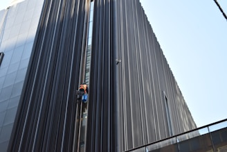 A person in bright safety gear is performing maintenance or cleaning work on a modern building's exterior. The structure features tall, vertical lines made of dark metallic or glass material, creating a striking visual contrast with the person.