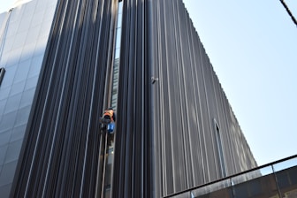 A person in bright safety gear is performing maintenance or cleaning work on a modern building's exterior. The structure features tall, vertical lines made of dark metallic or glass material, creating a striking visual contrast with the person.