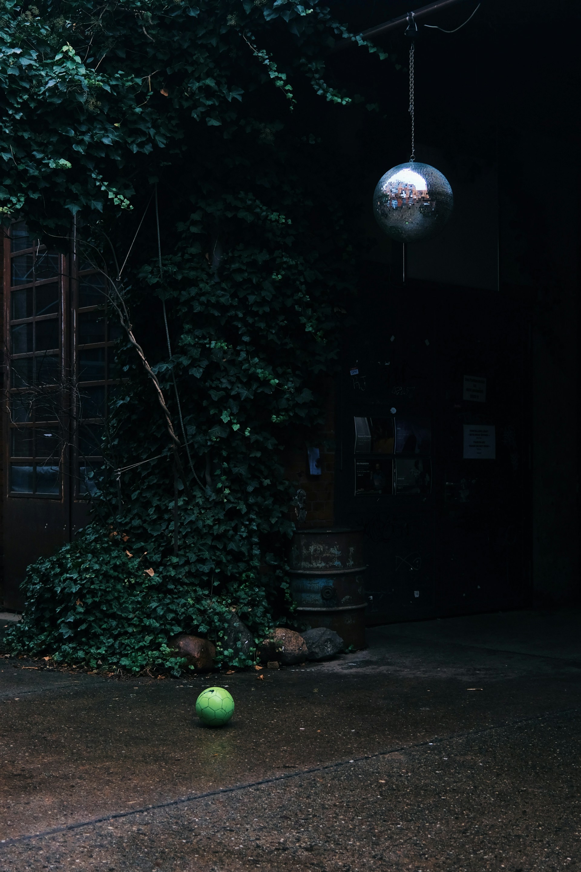 A dimly lit outdoor scene with a green soccer ball on a wet concrete surface beneath a hanging disco ball. The wall is covered with lush green ivy, and there is a wooden door with glass panes in the background.