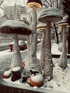 Large mushroom sculptures covered in snow are set in an urban environment with buildings in the background and cars parked along the street. The ground is blanketed with snow, and the streetlights create a cozy, warm glow under the mushroom caps.
