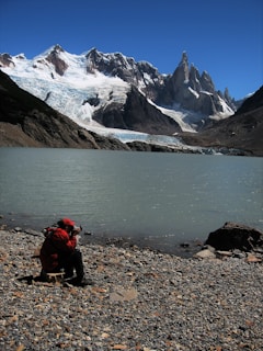 A person wearing a red jacket is seated on a rocky shoreline, taking a photograph of a scenic landscape that features a large glacier and jagged mountain peaks under a clear blue sky.