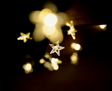 A collection of star-shaped string lights emitting a warm, soft glow against a dark background. The lights are slightly blurred in the distance, creating a dreamy and gentle atmosphere.