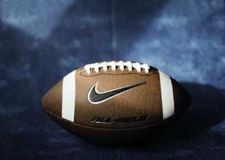 A well-lit American football rests on a textured blue surface. The football is brown with white laces and stripes, featuring a prominent Nike logo. The background is slightly blurred, creating a sense of focus on the football.