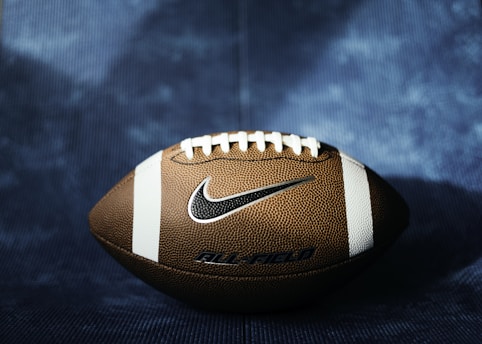 A well-lit American football rests on a textured blue surface. The football is brown with white laces and stripes, featuring a prominent Nike logo. The background is slightly blurred, creating a sense of focus on the football.