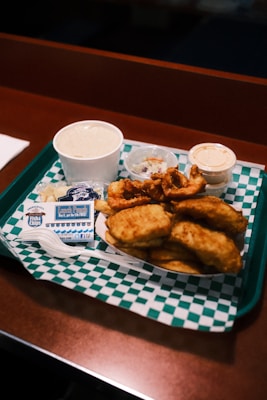 A food tray featuring a variety of fried seafood items is arranged on a table. The tray includes fried fish and calamari rings along with several dipping sauces in small containers. A small, closed seafood restaurant seals packet, a plastic fork, and a cup of clam chowder are also visible. The tray is lined with a green and white checkered paper.