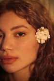 A close-up profile of a person's face, highlighting their features with soft lighting. The skin tone is warm and natural, with a focus on the eye and full lips. A white flower is placed behind the ear, contrasting with the person's curly, light brown hair.