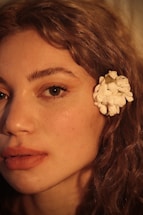 A close-up profile of a person's face, highlighting their features with soft lighting. The skin tone is warm and natural, with a focus on the eye and full lips. A white flower is placed behind the ear, contrasting with the person's curly, light brown hair.