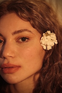 A close-up profile of a person's face, highlighting their features with soft lighting. The skin tone is warm and natural, with a focus on the eye and full lips. A white flower is placed behind the ear, contrasting with the person's curly, light brown hair.