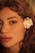 A close-up profile of a person's face, highlighting their features with soft lighting. The skin tone is warm and natural, with a focus on the eye and full lips. A white flower is placed behind the ear, contrasting with the person's curly, light brown hair.