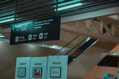 An interior of what appears to be a train or bus station, featuring a directional sign indicating locations like CBD Ciledug, Puri Beta, and more. The sign also points to facilities such as a prayer room, toilet, and ATM. There is an escalator and structural beams, along with panels displaying information or maps.