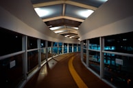 A modern, curved pedestrian walkway with large windows and a wooden floor. The ceiling features geometric patterns with inset lighting, creating a futuristic ambiance. The pathway includes a tactile guide path for the visually impaired.