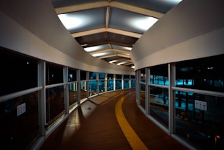 A modern, curved pedestrian walkway with large windows and a wooden floor. The ceiling features geometric patterns with inset lighting, creating a futuristic ambiance. The pathway includes a tactile guide path for the visually impaired.