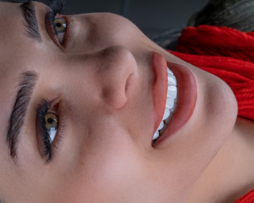 Close-up of a smiling person showing prominent white teeth, focusing on their eyes and eyebrows. The person is wearing a bright red scarf or garment, and the image conveys a sense of warmth and cheerfulness.