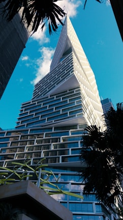 A modern skyscraper with an intricate, staggered design featuring numerous glass windows. The structure rises sharply against a clear blue sky, flanked by palm trees. The building's architecture includes unique, angular surfaces and projections.