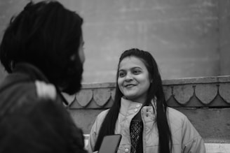 Two people are engaged in a conversation. One person is smiling, wearing a light-colored jacket, and has long hair. They are standing near a wall with decorative patterns.