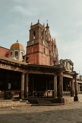 A historic Gothic-style church with a prominent clock tower stands in a sunny plaza. The architecture features intricate spires and stone columns, with a warm, golden hue cast by the sunlight.