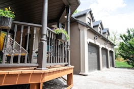 A modern house exterior featuring a porch with wooden railings and hanging flower baskets. Three garage doors are aligned on the side of the house. The structure has gabled roofs and decorative lighting fixtures. The surrounding area includes some greenery and a partially cloudy sky.