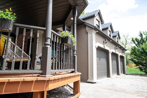 A modern house exterior featuring a porch with wooden railings and hanging flower baskets. Three garage doors are aligned on the side of the house. The structure has gabled roofs and decorative lighting fixtures. The surrounding area includes some greenery and a partially cloudy sky.