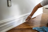 A person is painting the baseboard of a wall with a brush. The floor is made of polished wood and a blue cloth is on the floor nearby. The wall features a power outlet without a cover.