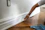 A person is painting the baseboard of a wall with a brush. The floor is made of polished wood and a blue cloth is on the floor nearby. The wall features a power outlet without a cover.