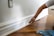 A person is painting the baseboard of a wall with a brush. The floor is made of polished wood and a blue cloth is on the floor nearby. The wall features a power outlet without a cover.