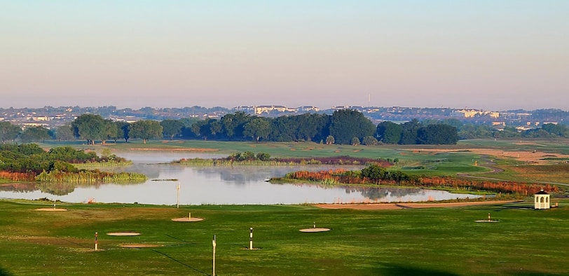 A serene landscape of a golf course is visible with a lake in the middle, surrounded by lush greenery and trees. The horizon features distant buildings under a clear sky. The foreground shows neatly manicured lawns with golf holes.