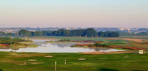 A serene landscape of a golf course is visible with a lake in the middle, surrounded by lush greenery and trees. The horizon features distant buildings under a clear sky. The foreground shows neatly manicured lawns with golf holes.