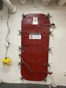 A heavy red door with metal fasteners and a sign indicating it is a controlled area for authorized and escorted personnel only. The door is set within a white wall and is surrounded by pipes. A yellow emergency light is mounted to the left of the door.