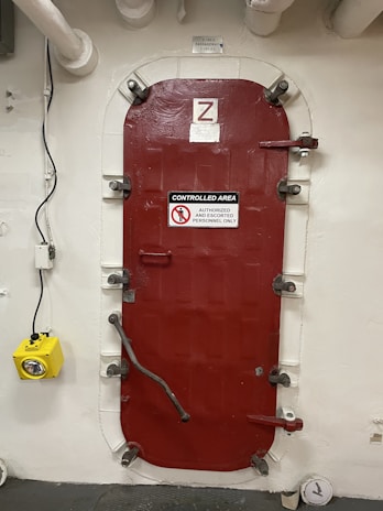 A heavy red door with metal fasteners and a sign indicating it is a controlled area for authorized and escorted personnel only. The door is set within a white wall and is surrounded by pipes. A yellow emergency light is mounted to the left of the door.