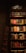 A dimly lit bookstore shelf stacked with a variety of books. The shelves contain books of different colors and sizes, organized in sections. Labels such as 'Çok Satanlar' (Bestsellers) and 'Yeni Çıkanlar' (New Arrivals) are visible. The surrounding ambiance is cozy with a brick wall partially visible to the left.