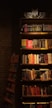A dimly lit bookstore shelf stacked with a variety of books. The shelves contain books of different colors and sizes, organized in sections. Labels such as '&Ccedil;ok Satanlar' (Bestsellers) and 'Yeni &Ccedil;ıkanlar' (New Arrivals) are visible. The surrounding ambiance is cozy with a brick wall partially visible to the left.