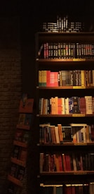 A dimly lit bookstore shelf stacked with a variety of books. The shelves contain books of different colors and sizes, organized in sections. Labels such as 'Çok Satanlar' (Bestsellers) and 'Yeni Çıkanlar' (New Arrivals) are visible. The surrounding ambiance is cozy with a brick wall partially visible to the left.