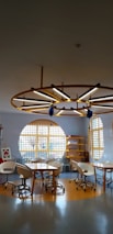 A modern, well-lit room with circular tables surrounded by white swivel chairs. The room features large round windows or wall decor with yellow grid patterns, complemented by shelves filled with books or materials. A unique ceiling structure with a circular wooden frame and integrated lighting adds a stylish touch.