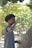 A person with curly hair is drinking from a green bottle while standing outdoors. The background features green leaves and trees, suggesting a natural environment.
