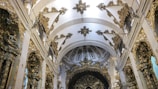 An ornately decorated interior of a historic building features intricate golden detailing and sculptural elements on the walls and ceilings. The vaulted ceiling is adorned with elaborate patterns and decorative motifs, creating a sense of grandeur and elegance.