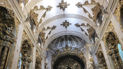An ornately decorated interior of a historic building features intricate golden detailing and sculptural elements on the walls and ceilings. The vaulted ceiling is adorned with elaborate patterns and decorative motifs, creating a sense of grandeur and elegance.