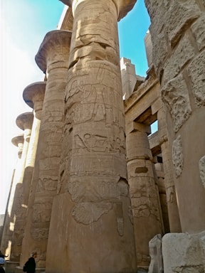 Massive ancient stone columns intricately carved with hieroglyphs and symbols, suggesting an ancient Egyptian temple or structure. The surrounding architecture is grand and imposing, with a bright blue sky visible above.