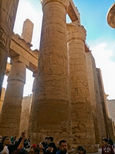 Massive ancient stone columns with intricate hieroglyphic carvings dominate the scene. Several tourists stand at the base, looking up and taking pictures. The architecture exudes a sense of history and grandeur, characteristic of ancient Egyptian temples.