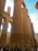 Massive ancient stone columns with intricate hieroglyphic carvings dominate the scene. Several tourists stand at the base, looking up and taking pictures. The architecture exudes a sense of history and grandeur, characteristic of ancient Egyptian temples.