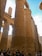 Massive ancient stone columns with intricate hieroglyphic carvings dominate the scene. Several tourists stand at the base, looking up and taking pictures. The architecture exudes a sense of history and grandeur, characteristic of ancient Egyptian temples.