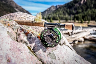 A close-up of a fly fishing rod and reel resting on a rugged rock by a river. The rod features a detailed fly lure above the reel. The background includes a blurred view of a forested mountain landscape under a blue sky.