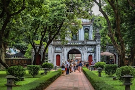 A historical gate with an ornate design stands along a pathway, surrounded by lush green trees and manicured bushes. A group of people is gathered near the entrance, suggesting a tourist attraction or cultural site. Traditional stone lanterns line the path, enhancing the serene and historic atmosphere.