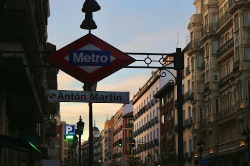 A city street with a prominent metro sign named 'Antón Martin'. The scene includes surrounding buildings with classic architecture, street lamps, and signs indicating a parking area. The sky has a soft sunset hue.